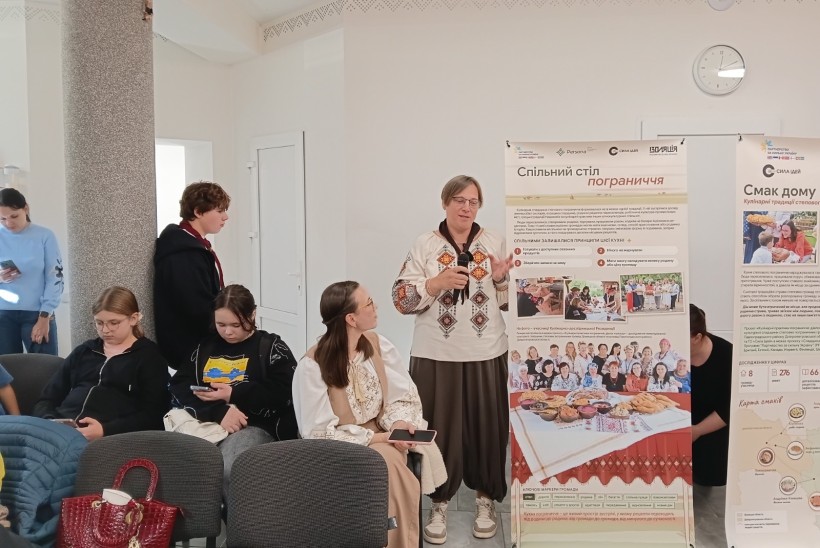 Ведуча у вишиванці виступає з мікрофоном біля інформаційного стенда про кулінарні традиції перед слухачами.