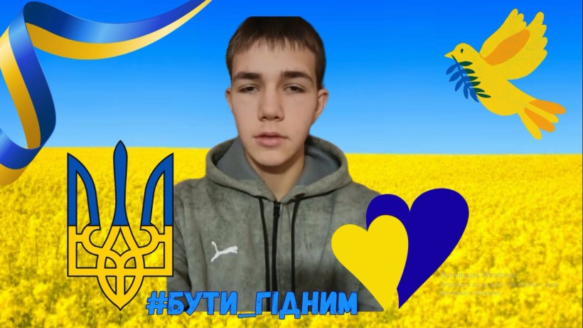 Хлопець, учасник флешмобу, у сірій толстовці на тлі жовто-блакитного прапора та поля із зображенням тризуба, жовтого й синього сердець