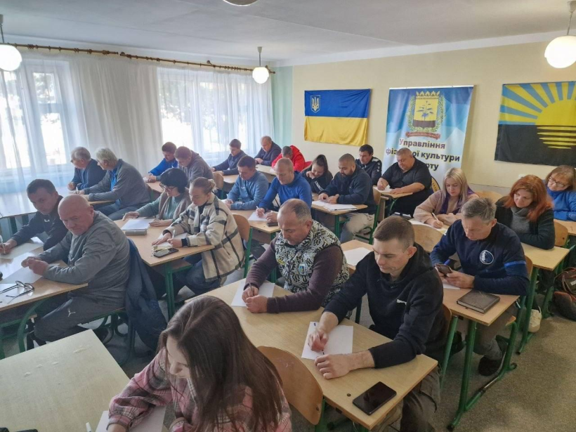 Представники управління фізичної культури та спорту сидять за партою в кімнаті з українськими прапорами та пишуть диктант.