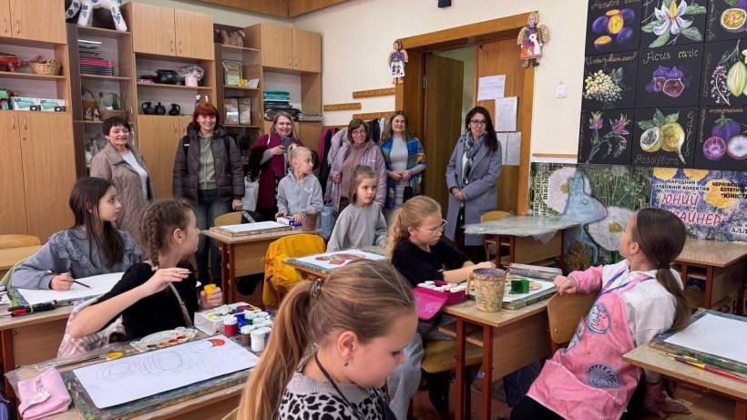 У художній майстерні кілька дівчат малюють за партами.  Біля дверей стоїть група жінок — гостей із Добропілля.