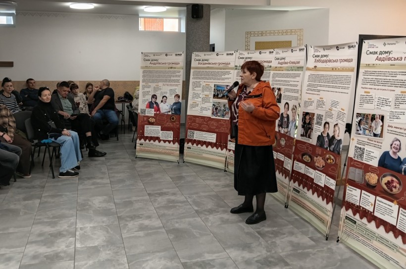 Ведуча говорить у мікрофон біля ряду виставочних банерів.