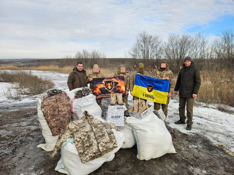 Дія по серед поля. На білих мішках лежить нова форма для військових. Поруч волонтери та представники громади.
