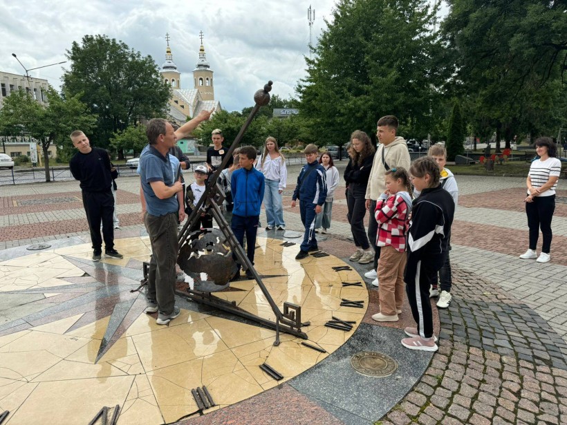 Діти біля сонячного годинника. Екскурсовод рукою показує напрямок сонячного світла. Діти уважно слухають.