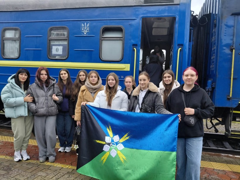 Група молодих людей і жінка стоять, посміхаючись, перед синім поїздом, тримаючи в руках кольоровий прапор. Настрій веселий і жвавий.