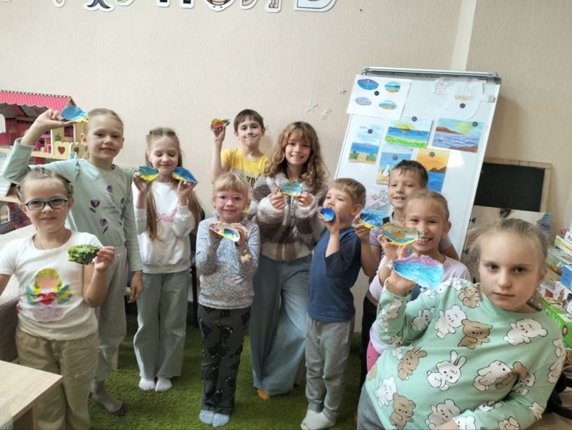 Група усміхнених дітей з гордістю демонструє свої барвисті малюнки в класі. На дошці в задньому плані виставлено ще більше художніх робіт.