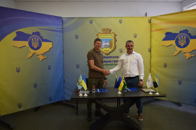 Голови громад тиснуть друг другу руки. Стоять біля столу, на якому лежать документи та стоять прапори громад.