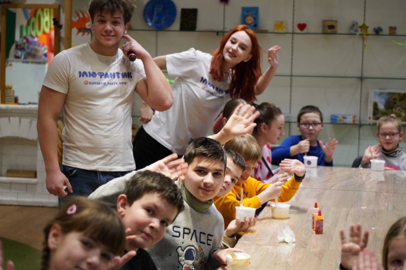 Група дітей та двоє аніматорів у білих футболках махають руками, сидячи за довгим столом.