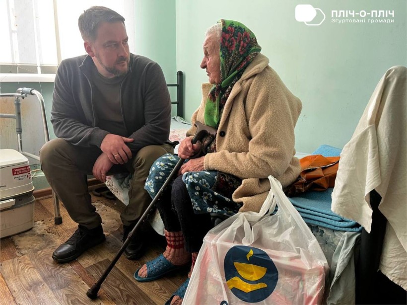Заступник начальника Часовоярської міської військової адміністрації Руслан Прийменко сидить поруч біля жінки похилого віку, спілкується, поруч стоїть пакет гуманітарної допомоги.