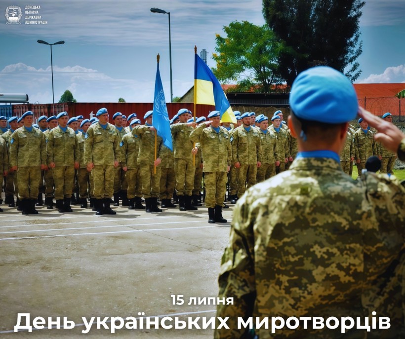 Солдати в камуфляжі та блакитних беретах стоять у строю, один віддає честь. Вони тримають прапори України та ООН під частково хмарним небом.