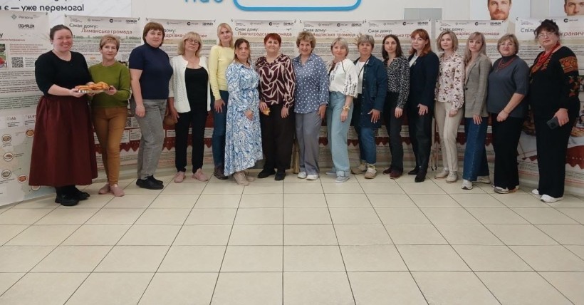 Учасниці заходу позують для фото в ряд біля виставочних банерів, одна з них тримає піднос із випічкою