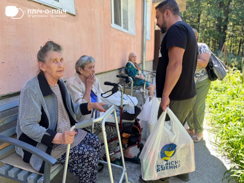 Жінки похилого віку сидять на вулиці біля будівлі, поруч з ними ходунки. Заступник начальника Часовоярської громади стоїть біля них з пакетом гуманітарної допомоги. Спілкуються.