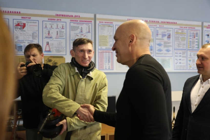 Вадим Філашкін тисне руку педагогу в робочому костюмі. Позаду фоторепортер.