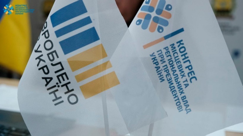 Два білих прапори з синьо-жовтими геометричними візерунками та текстом. Зображення передає почуття національної та організаційної гордості.