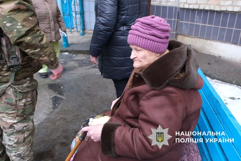 Жінка похилого віку сидить на лавці, біля неї поліцейські,які допомагають з евакуацією.