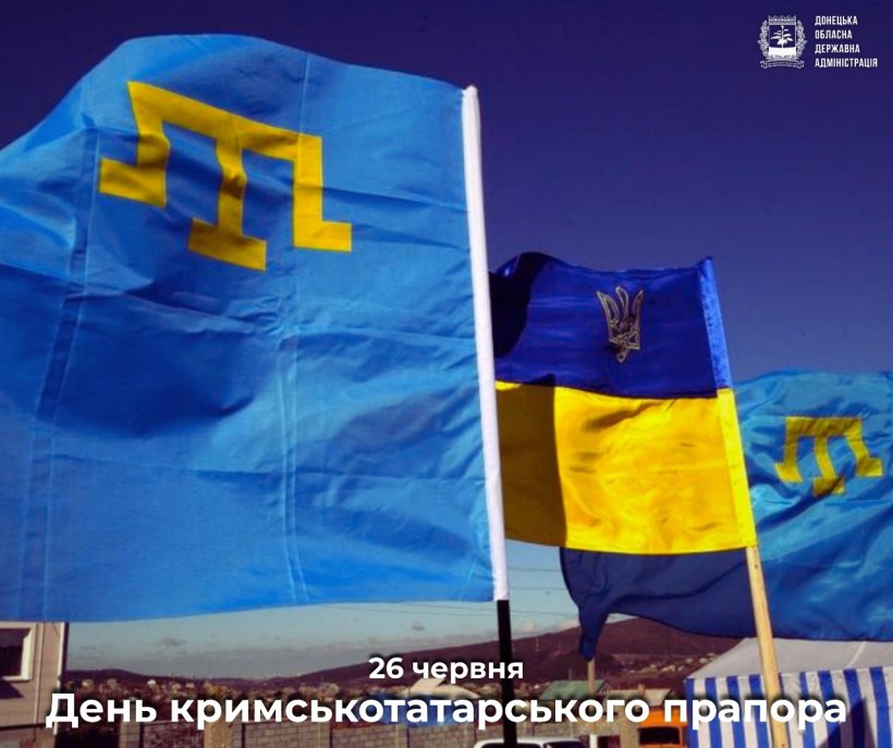 Яскраво-синій прапор кримських татар з жовтою емблемою майорить поруч з українськими прапорами під чистим небом, відзначаючи День прапора кримських татар.