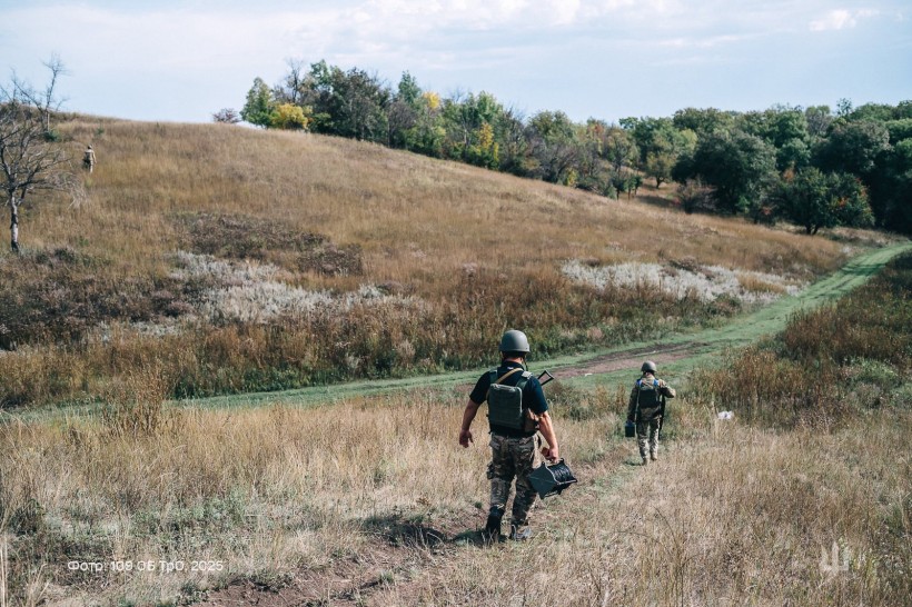 Двоє захисників ідуть стежкою. В руках тримають знаряддя. Попереду посадка.