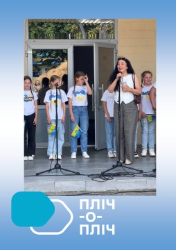 Діти Іркліївської сільської  громади вітають дітей з Шахівської сільської громади. Дівчата стоять с прапорцями України, одягнені у джинси та білі футболки з мапою країни. Ведуча говорить у мікрофон.