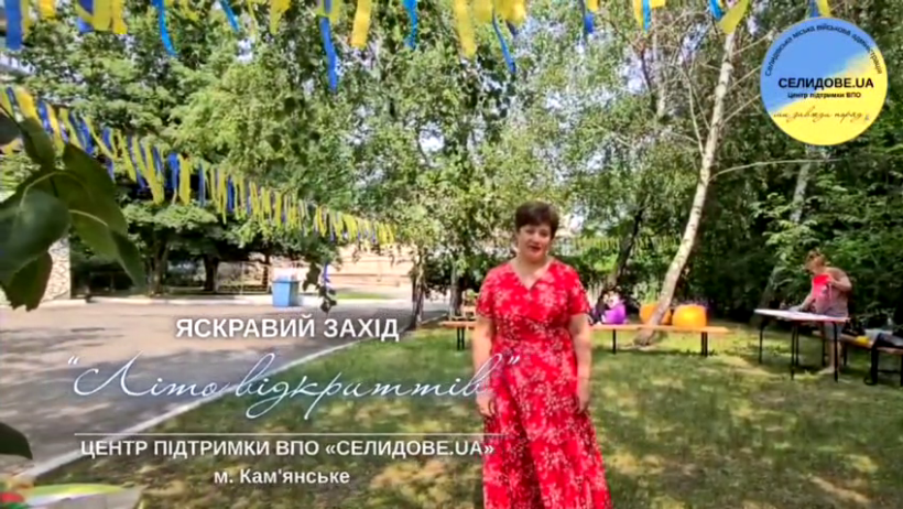 Урочисте відкриття табору "Літо відкриттів" у Кам'янському для дітей з Селидівської громади. Територія прикрашена патріотичними стрічками, у центрі-жінка у червоній сукні на фоні арт-зони