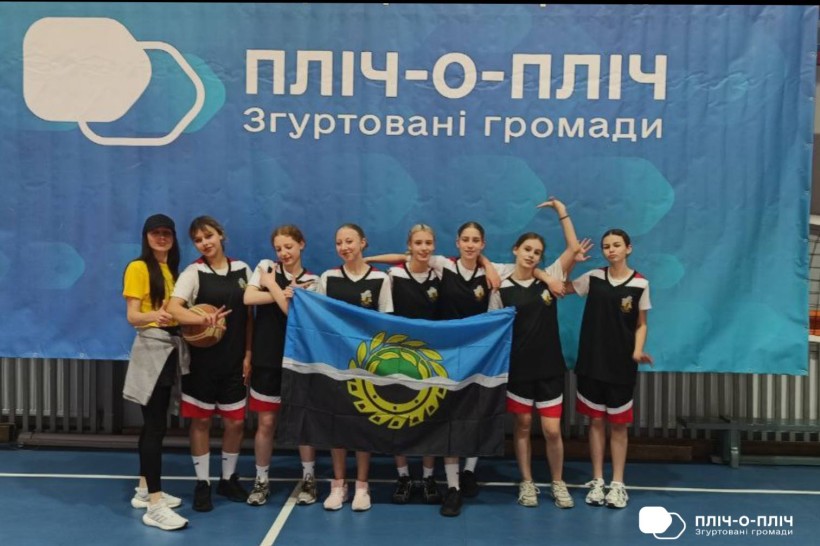 Група дівчат-спортсменок з Білозерської громади тримає прапор громади та позує на фоні банеру проєкту "Пліч-о-пліч"