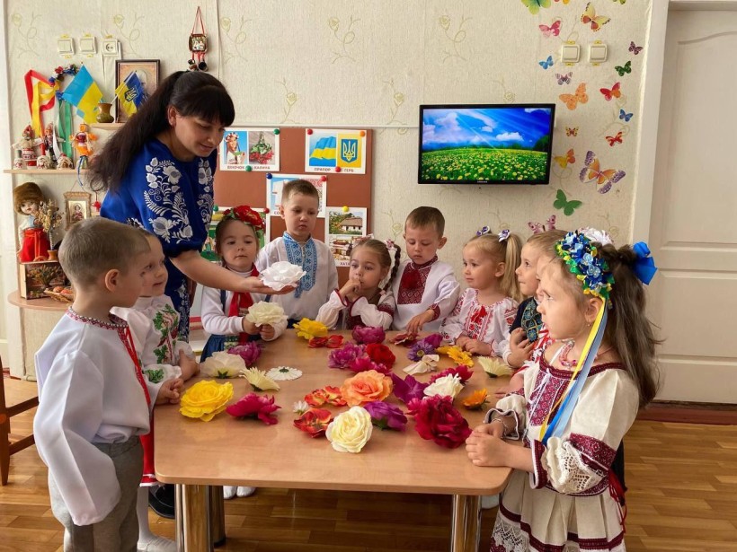 Практикантка педколеджу навчає дошкільнят українським традиціям, створюючи квіти. Діти у вишиванках, навколо українська символіка.