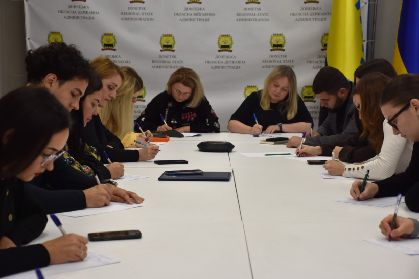Службовці обладміністрації за столом зосереджено пишуть диктант у залі з прапорами України та символікою Донецької області.