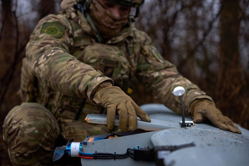 Солдат у камуфляжі налаштовує невеликий дрон на відкритому повітрі. Місце дії — лісиста місцевість, що наводить на думку про тактичну або розвідувальну місію. Атмосфера зосереджена.