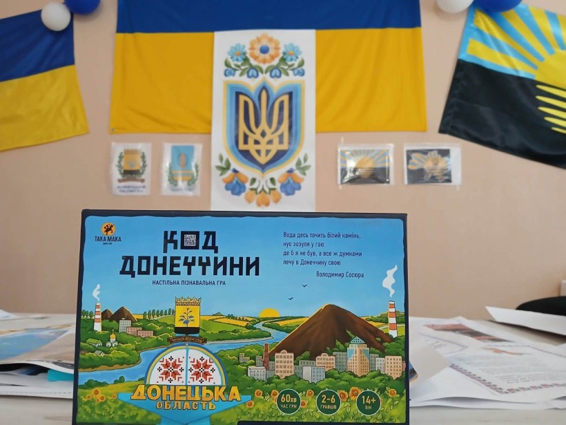 Гра "Код Донеччини" у коробці на столі, поруч з яким висять прапори України та Донецької області