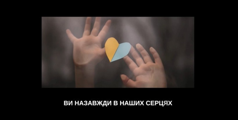 Руки дитини тримають паперове серце у кольорах прапора України, знизу напис «Ви назавжди в наших серцях»