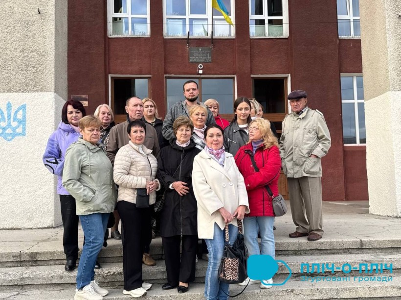 Група людей різного віку стоїть на сходах перед червоною будівлею з великими вікнами. Над входом висить український прапор. Вони виглядають серйозними та уважними.