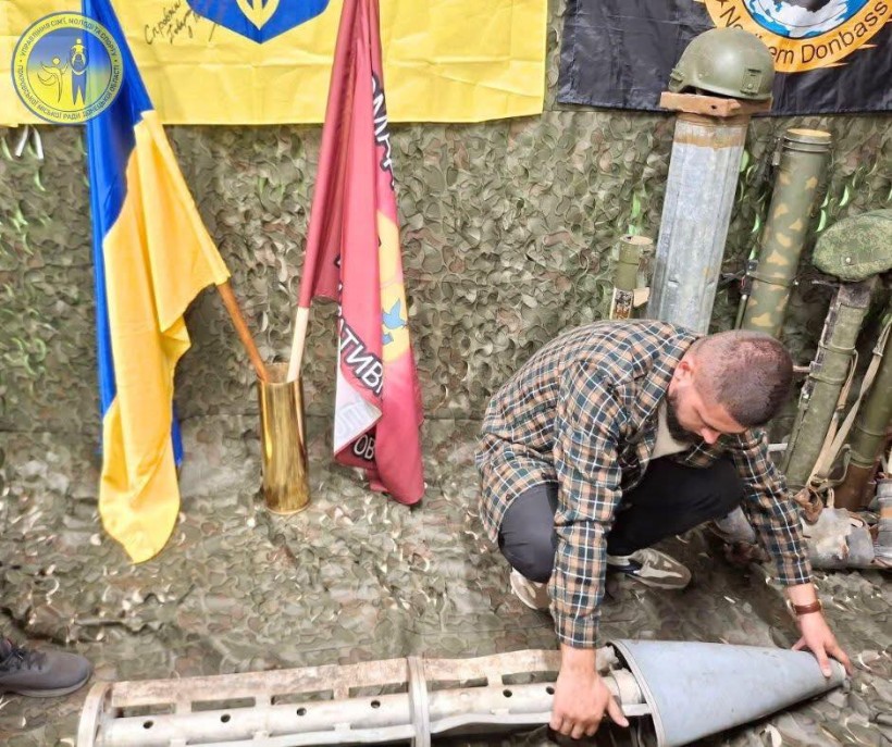 На полу лежить уламок снаряду, хлопець уважно розглядає. Позаду нього прапори бригад та захисна сітка зеленого кольору та тубуси.