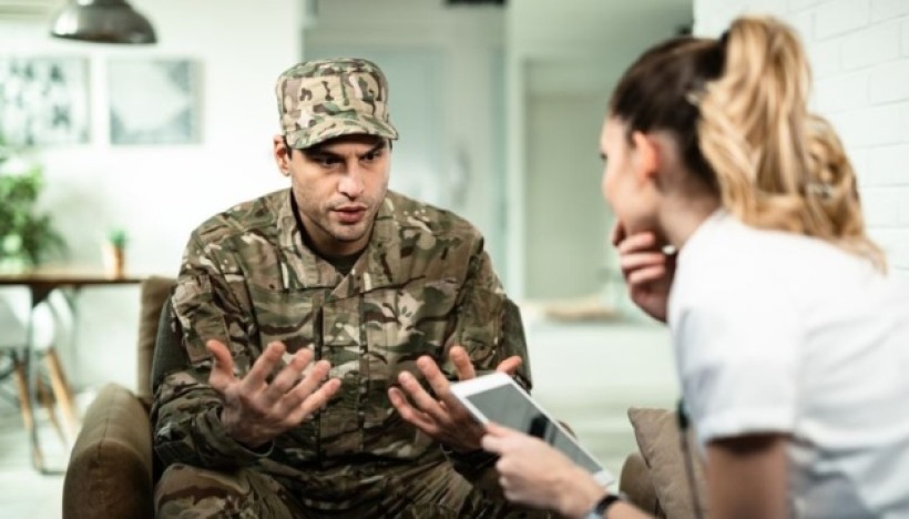 Людина у військовій формі сидить на дивані, жестикулюючи під час розмови з жінкою, яка тримає планшет. Обстановка невимушена, розмова ведеться у невимушеній манері.