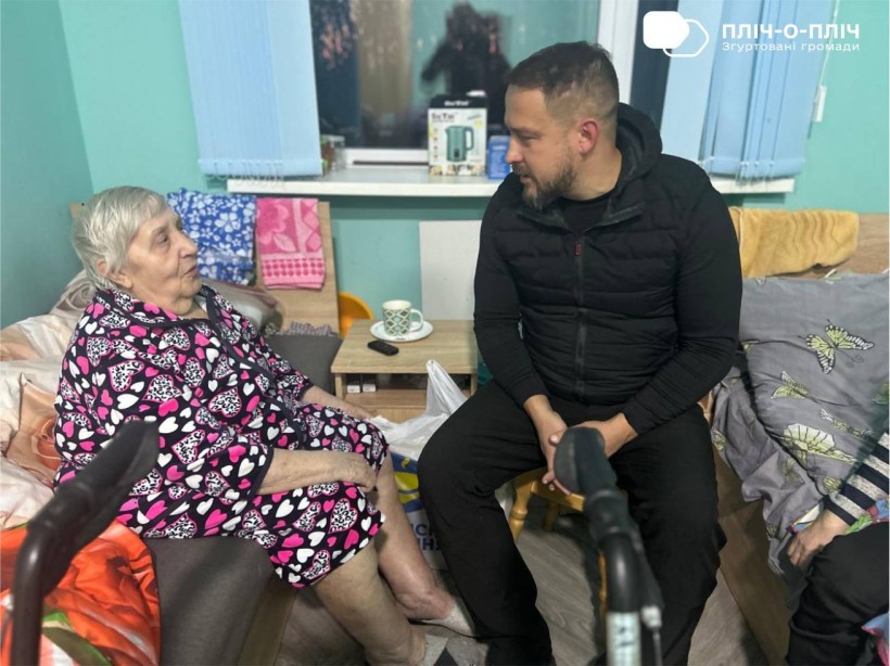 Заступник начальника Часовоярської міської військової адміністрації Руслан Прийменко сидить біля жінки похилого віку, спілкується та слухає.