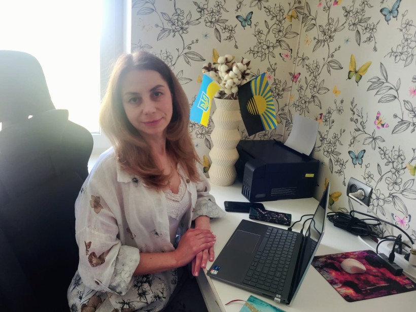 Жінка з русявим волоссям у світлій сорочці сидить за робочим столом біля вікна, на фоні шпалер із квітковим принтом та прапорів України й Донеччини.