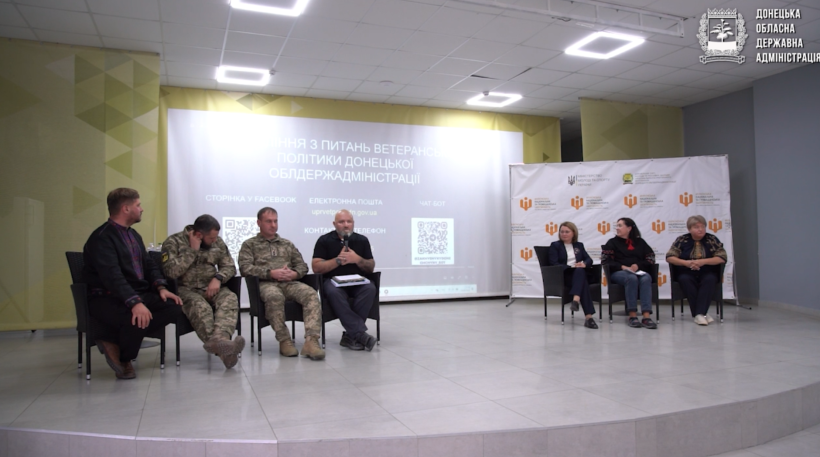 Сім осіб сидять на сцені під час панельної дискусії, двоє з них у військовій формі. За ними розташовані екран для презентацій та банери організації.