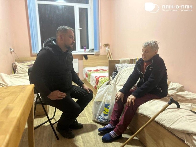 Жінка похилого віку,сидить на кроваті поруч дерев'яна палиця, напроти неї сидить на стільці Заступник начальника Часовоярської міської військової адміністрації Руслан Прийменко, який передав пакет з гуманітарною допомогою.