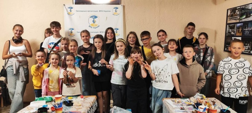 A group of smiling children and teens pose indoors in front of a banner. They are gathered around tables with art supplies, exuding joy and creativity.