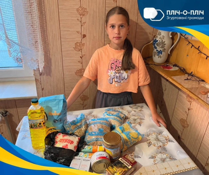 Дівчина стоїть біля столу, де викладена продуктова допомога