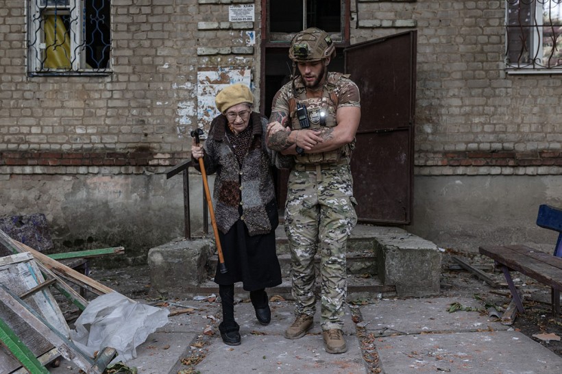 Літня жінка з палицею виходить з під'їзду будинку, поліцейський тримає її під руку.
