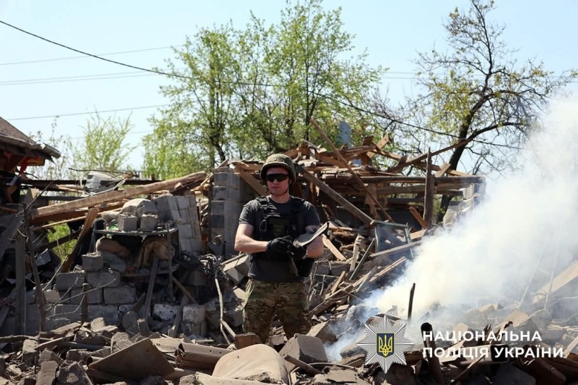 Поліцейський у повній екіпіровці, тримає уламок снаряда. На задньому плані дим та уламки.