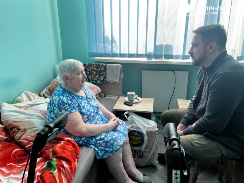 Заступник начальника Часовоярської міської військової адміністрації Руслан Прийменко спілкується з жінкою похилого віку яка сидить на кроваті.