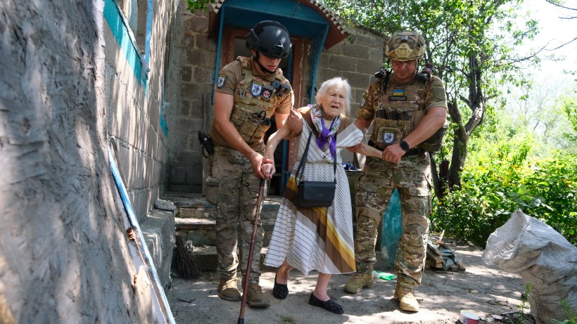 Солдати в камуфляжі та шоломах допомагають літній жінці з палицею біля кам'яного будинку. Їх оточує пишна зелень, що передає відчуття терміновості та турботи.