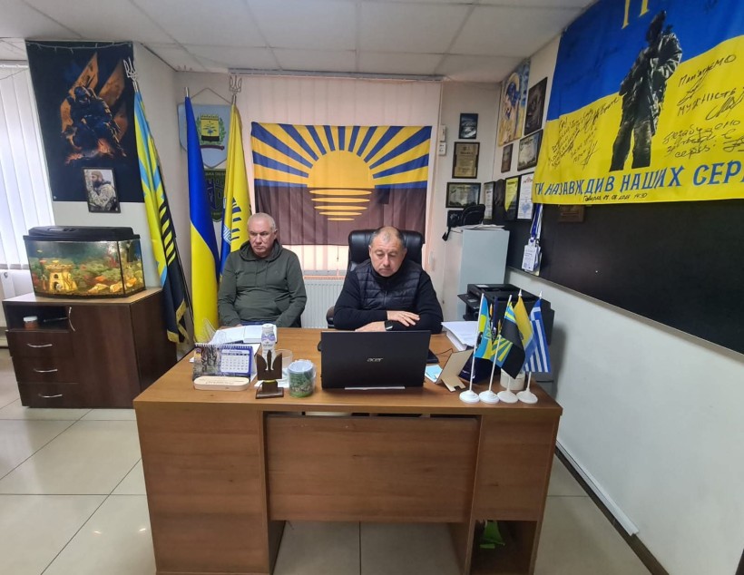 Двоє чоловіків сидять в офісі з патріотичними прапорами, фотографіями в рамках і ноутбуком на столі. Атмосфера офіційна і зосереджена.