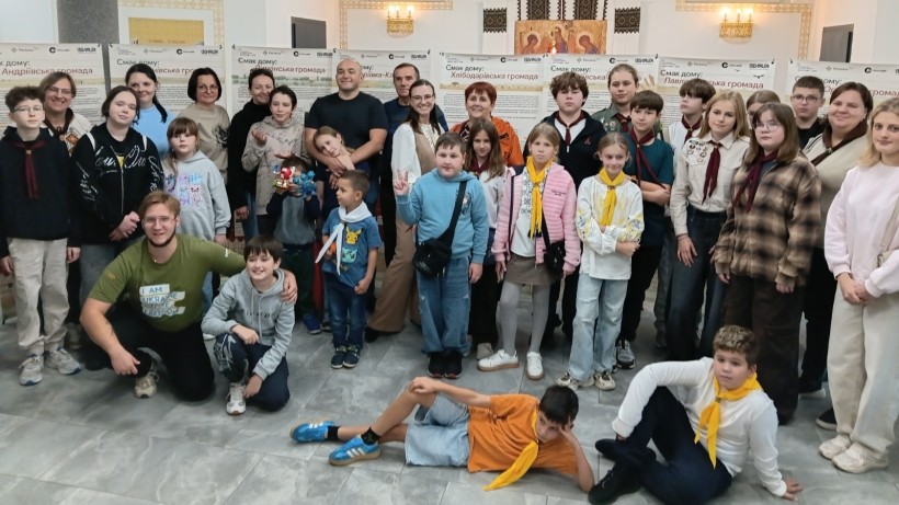 Спільне групове фото дітей та дорослих у залі на тлі виставочних стендів.