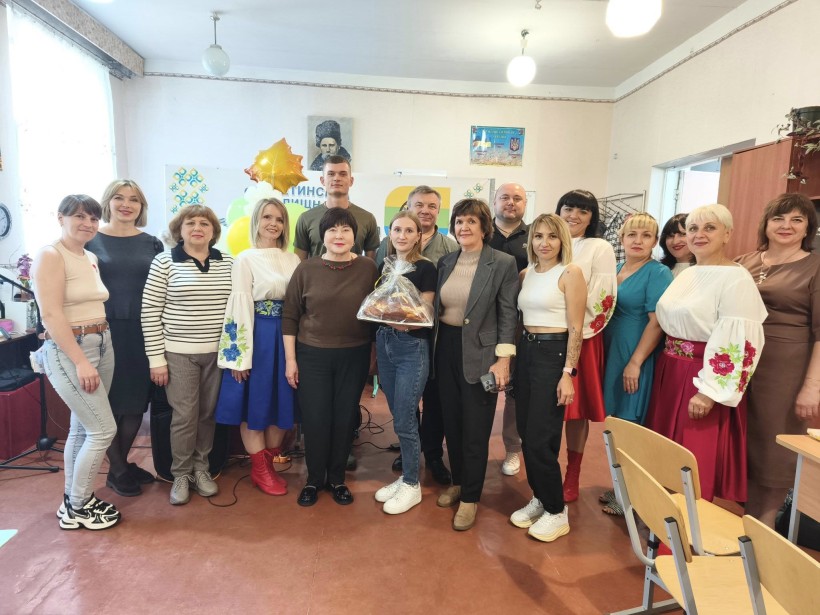 Спільне фото. Жінки та чоловіки стоять у приміщенні, молода жінка в руках тримає паляницю.