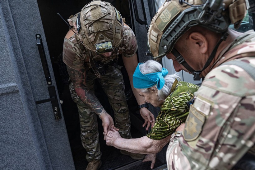 Двоє поліцейських допомагають сісти у автомобіль літній жінці. Жінка однією рукою тримається за сходи авто.
