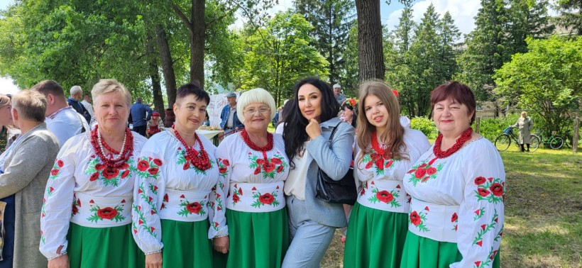 Учасниці пісенного фестивалю. У вишиванках та зелених спідницях, на шиях червоні намиста. Поруч жінка у строгому діловому костюмі.