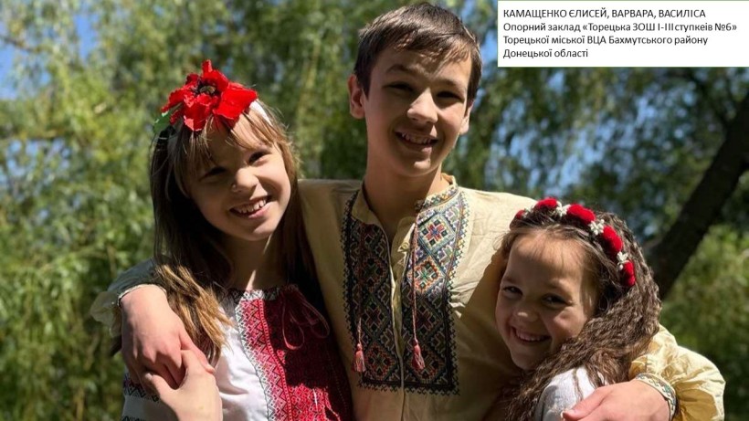 Троє усміхнених дітей у вишиванках обіймаються на фоні зелених дерев.