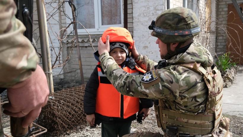 Солдат у камуфляжі допомагає маленькій дитині в помаранчевому шоломі та жилеті. Сцена відбувається на відкритому повітрі біля цегляної будівлі, що створює відчуття захисту та турботи.