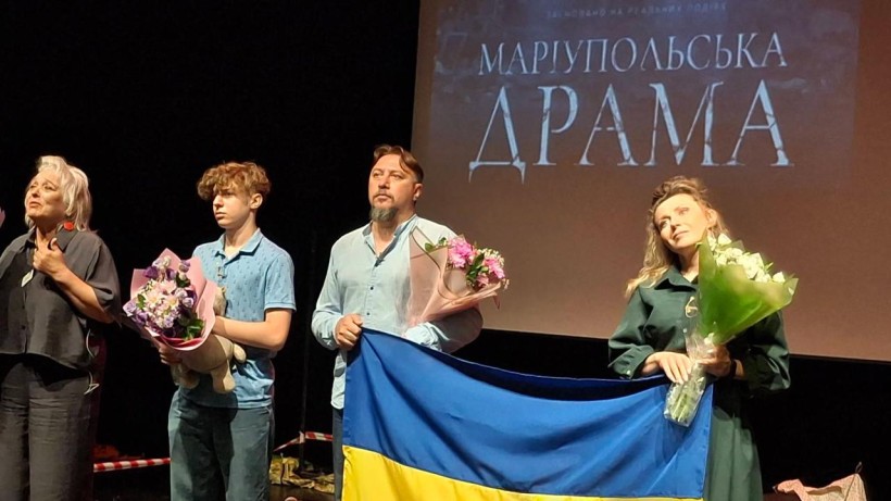 Актори вистави «Маріупольська драма» на сцені тримають прапор України та квіти на тлі екрана з назвою спектаклю.