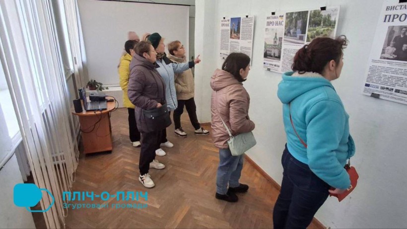 Учасники виставки стоять біля фотографій, одна з учасниць щось показує рукою.